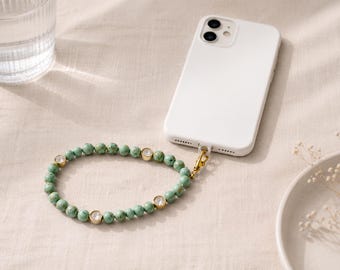 Könnte beinhalten: Ein Handy-Armband mit runden, marmorierten grünen Perlen, die mit vier goldfarbenen Elementen mit klaren, runden Kristallen durchsetzt sind. Die Perlenschlaufe ist mit einem goldfarbenen Karabiner und einer transparenten Schnur am weißen Smartphone befestigt.