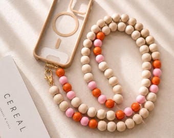 Könnte beinhalten: Eine transparente Handyhülle mit beigem Rahmen und magnetischem Ring, verbunden mit einem langen Umhängeband aus runden Holzperlen in Naturbeige, leuchtendem Orange und zartem Rosa, ausgestattet mit goldenen Karabinerverschlüssen.