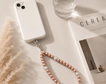 Könnte beinhalten: Eine weiße Smartphone-Hülle mit einer Handschlaufe aus runden, metallisch roségoldenen Perlen, die mit vier ovalen, weißen, perlmuttartigen Akzenten durchsetzt sind. Die Schlaufe hat einen silberfarbenen Karabinerverschluss aus Metall.