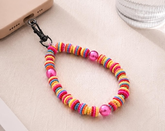 Könnte beinhalten: Ein Handy-Armband mit einem sich wiederholenden Muster aus kleinen, bunten, scheibenförmigen Perlen in Gelb, Blau, Orange und Pink, durchsetzt mit drei größeren, glatten, metallisch pinkfarbenen Kugeln, befestigt an einem schwarzen Metallkarabiner.