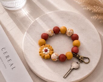 Könnte beinhalten: Ein kreisförmiges Schlüsselanhänger-Armband mit einer Mischung aus senfgelben, terrakotta-roten und floral gemusterten Perlen. Eine große senfgelbe blumenförmige Perle ist am Band befestigt, das einen bronzefarbenen Karabinerhaken und einen Schlüssel enthält.