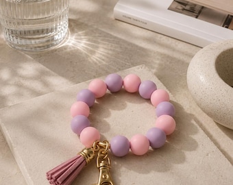 Könnte beinhalten: Ein kreisförmiges Schlüsselanhänger-Armband mit abwechselnd matten rosa und violetten Silikonperlen. Das Armband verfügt über einen goldfarbenen Karabinerverschluss aus Metall und eine passende rosa Quaste aus Kunstleder an einem goldenen Ring.