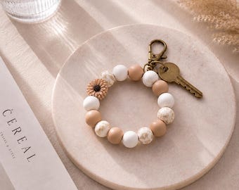 Könnte beinhalten: Ein kreisförmiger Schlüsselanhänger aus glatten, runden Perlen in Weiß-, Beige- und Brauntönen, der eine kleine, texturierte Perle in Sonnenblumenform enthält. Der Schlüsselanhänger verfügt über einen bronzefarbenen Karabinerhaken und einen Metallschlüssel.