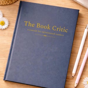 May include: A navy blue journal titled "The Book Critic" with gold lettering, next to two pencils and a cup of coffee. The journal is on a wooden surface, with daisies and pencils in a small white bowl.