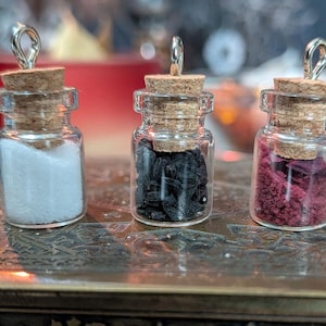 Könnte beinhalten: Drei kleine Glasflaschen mit Korken und silbernen Schlaufen. Die Flaschen enthalten weiße, schwarze und rote granulare Substanzen. Die Flaschen werden auf einer dekorativen Metalloberfläche ausgestellt.