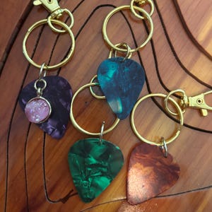 Puede incluir: Llaveros con colgantes de púas de guitarra en varios colores, incluyendo morado, verde y naranja. Cada púa está unida a un llavero y cierre de color dorado. Un llavero presenta un charm rosa brillante.