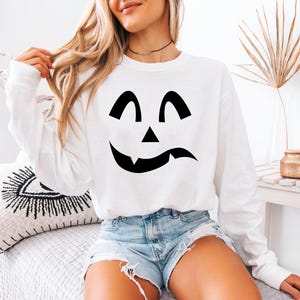 A woman wearing a white sweatshirt with a black jack-o-lantern design, sitting on a bed with a white pillow.