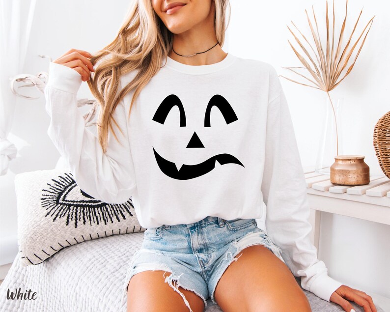 A woman wearing a white sweatshirt with a black jack-o-lantern design, sitting on a bed with a white pillow.