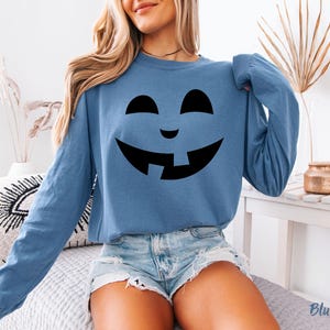 A woman wearing a blue long-sleeved shirt with a black graphic of a smiling jack-o-lantern on it, paired with denim shorts.