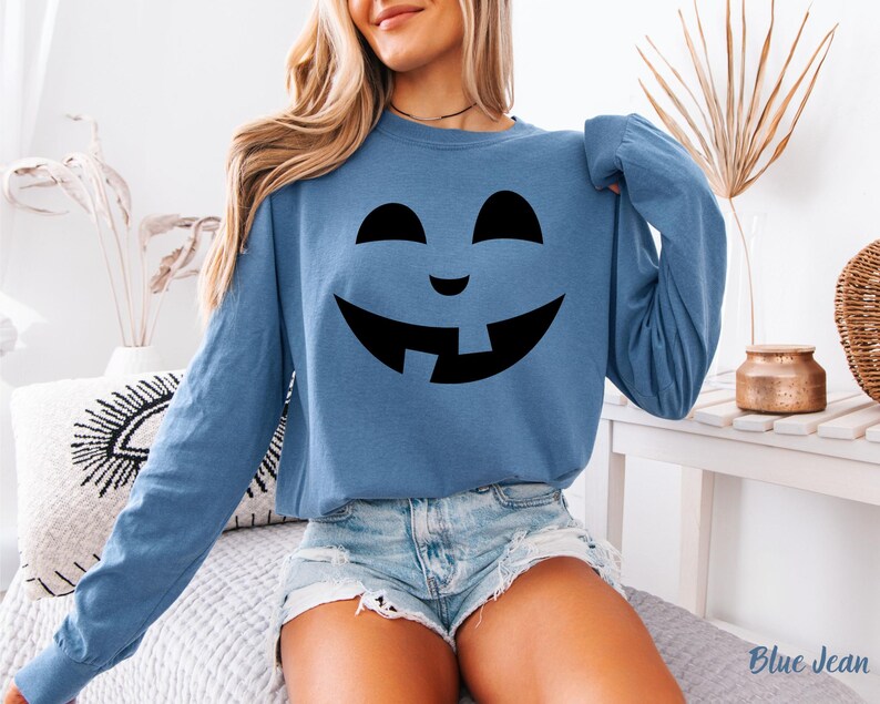 A woman wearing a blue long-sleeved shirt with a black graphic of a smiling jack-o-lantern on it, paired with denim shorts.