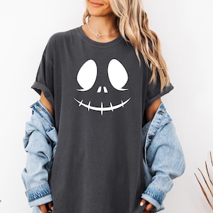 A woman wearing a black t-shirt with a white graphic of a smiling face and a denim jacket, standing against a white background.
