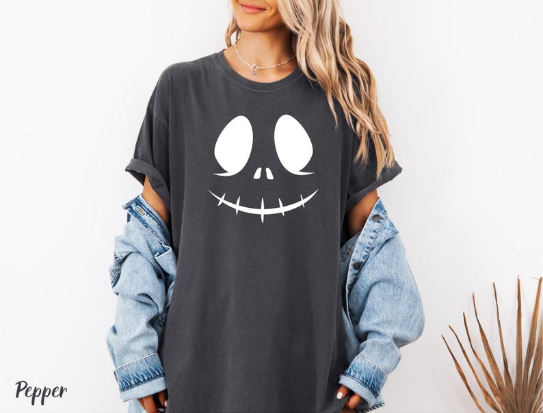 A woman wearing a black t-shirt with a white graphic of a smiling face and a denim jacket, standing against a white background.
