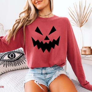 A woman wearing a red sweatshirt with a black jack-o-lantern graphic, denim shorts, and a necklace is sitting on a bed.