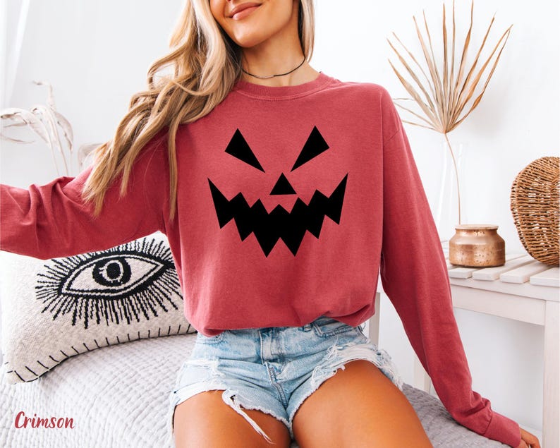 A woman wearing a red sweatshirt with a black jack-o-lantern graphic, denim shorts, and a necklace is sitting on a bed.