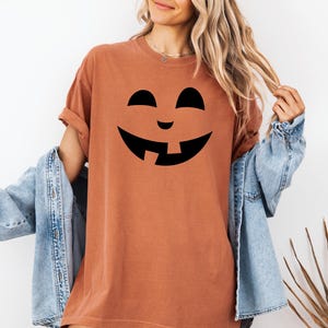 A woman wearing a t-shirt with a pumpkin face design and a denim jacket stands in front of a white wall.