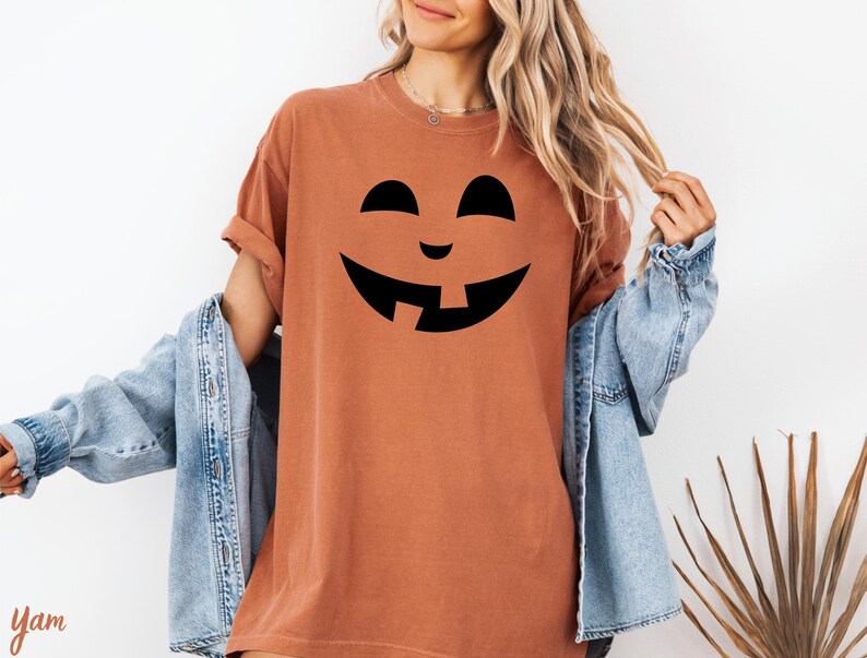 A woman wearing a t-shirt with a pumpkin face design and a denim jacket stands in front of a white wall.
