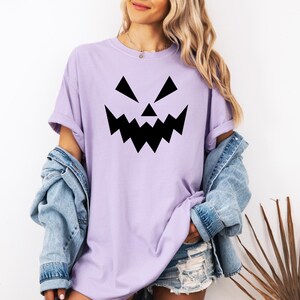 A woman wearing a purple t-shirt with a black jack-o-lantern graphic, a denim jacket, and denim shorts stands in front of a plant.