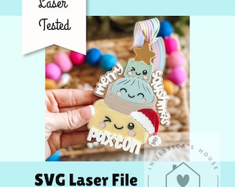 May include: A multi-layered ornament featuring a stack of three cute characters: a yellow square with a Santa hat, a light blue dumpling, and a small green square topped with a gold star. The text reads merry squishmas and paxton.