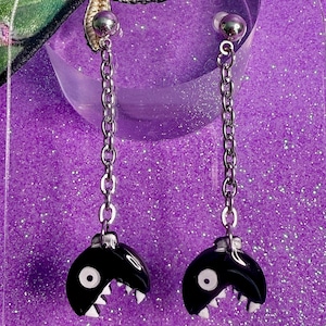 May include: A pair of black and white earrings with a chain design. The earrings feature a black, rounded shape with a white mouth and teeth design. The earrings are suspended from a silver chain and a silver ball stud.