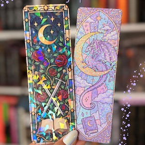 May include: Two rectangular holographic bookmarks with the text Holographic Bookmark at the top. The left bookmark features a crescent moon, red roses, two crossed swords, and an open book. The right bookmark displays a dragon, a crescent moon, and a book.