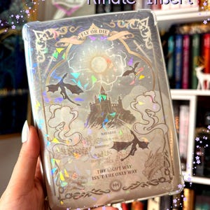 May include: A clear Kindle insert featuring a holographic design with a central castle, flying dragons, and the text "FLY OR DIE", "NAVARRE", "BASGIATH", and "THE RIGHT WAY ISN'T THE ONLY WAY". The graphic includes ornate gold-toned borders.