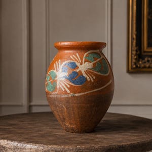 May include: A terracotta vase with a wide rim and a rounded body. The upper portion features hand-painted floral motifs in blue, green, and white, while the lower section remains a solid, unadorned dark brown clay with a textured, weathered finish.