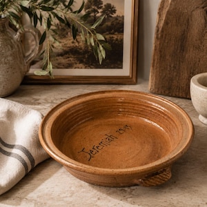 May include: A round, shallow, terracotta-colored ceramic baking dish with two small side handles. The interior base features the hand-etched text Jeremiah 29:11 in a dark, cursive script. The dish has a speckled, earthy glaze and a ridged rim.