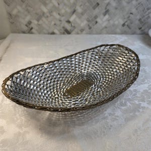 Puede incluir: Una cesta de forma ovalada con un interior de metal tejido plateado y un borde de alambre en tono bronce oscuro. La base de la cesta muestra un patr&oacute;n de rejilla rectangular hecho de finas tiras de metal color bronce oscuro en el centro.