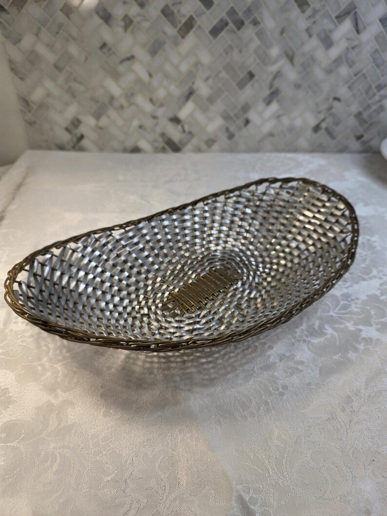 Puede incluir: Una cesta de forma ovalada con un interior de metal tejido plateado y un borde de alambre en tono bronce oscuro. La base de la cesta muestra un patr&oacute;n de rejilla rectangular hecho de finas tiras de metal color bronce oscuro en el centro.