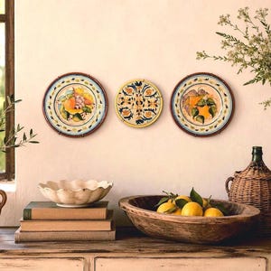 May include: Three circular ceramic wall plates featuring hand-painted designs of citrus fruits, grapes, and intricate floral patterns in shades of yellow, blue, and green, displayed horizontally on a wall above a wooden surface with various items.
