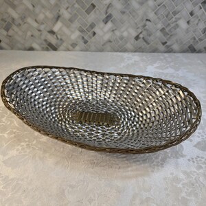 Puede incluir: Una cesta de forma ovalada con un cuerpo de metal plateado trenzado y un panel central distintivo de color bronce. El borde est&aacute; construido con un alambre fino de tono bronce oscuro, creando un contorno estructurado que enmarca el tejido plateado.