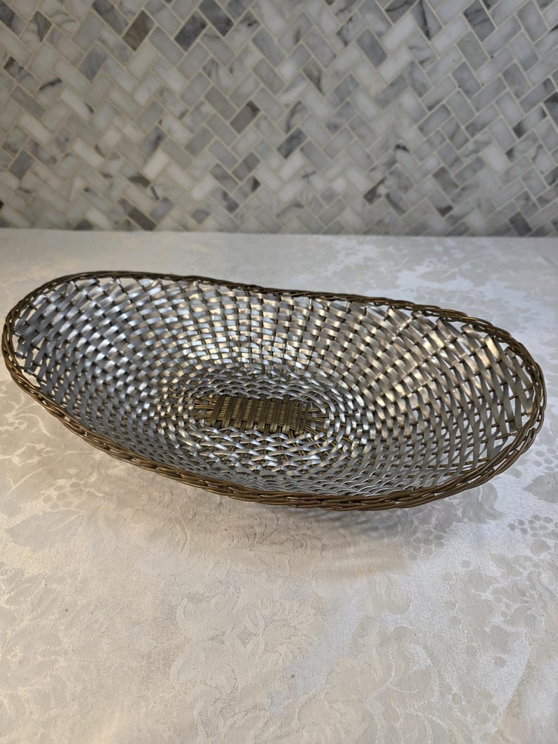 Puede incluir: Una cesta de forma ovalada con un cuerpo de metal plateado trenzado y un panel central distintivo de color bronce. El borde est&aacute; construido con un alambre fino de tono bronce oscuro, creando un contorno estructurado que enmarca el tejido plateado.