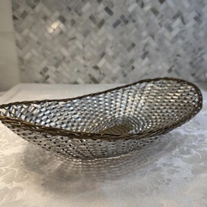 Puede incluir: Una cesta de forma ovalada con un cuerpo de metal plateado tejido y un borde de tono bronce oscuro. La estructura est&aacute; compuesta por finas tiras de metal entrelazadas que forman un patr&oacute;n de celos&iacute;a continuo creando una forma alargada.