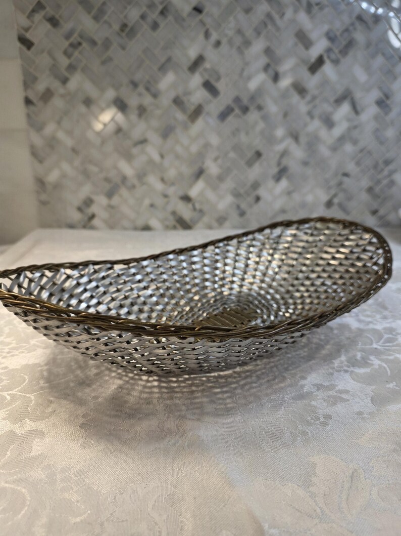 Puede incluir: Una cesta de forma ovalada con un cuerpo de metal plateado tejido y un borde de tono bronce oscuro. La estructura est&aacute; compuesta por finas tiras de metal entrelazadas que forman un patr&oacute;n de celos&iacute;a continuo creando una forma alargada.