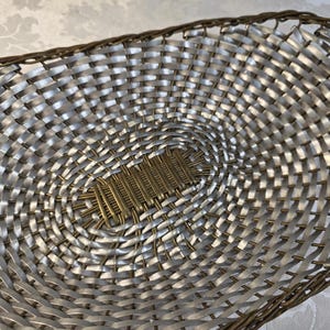 Puede incluir: Una cesta de forma ovalada con un patr&oacute;n trenzado de tiras plateadas met&aacute;licas y alambre fino de color bronce. La base central consiste en una secci&oacute;n rectangular de alambre de bronce tejido, mientras que el borde est&aacute; acabado en alambre.