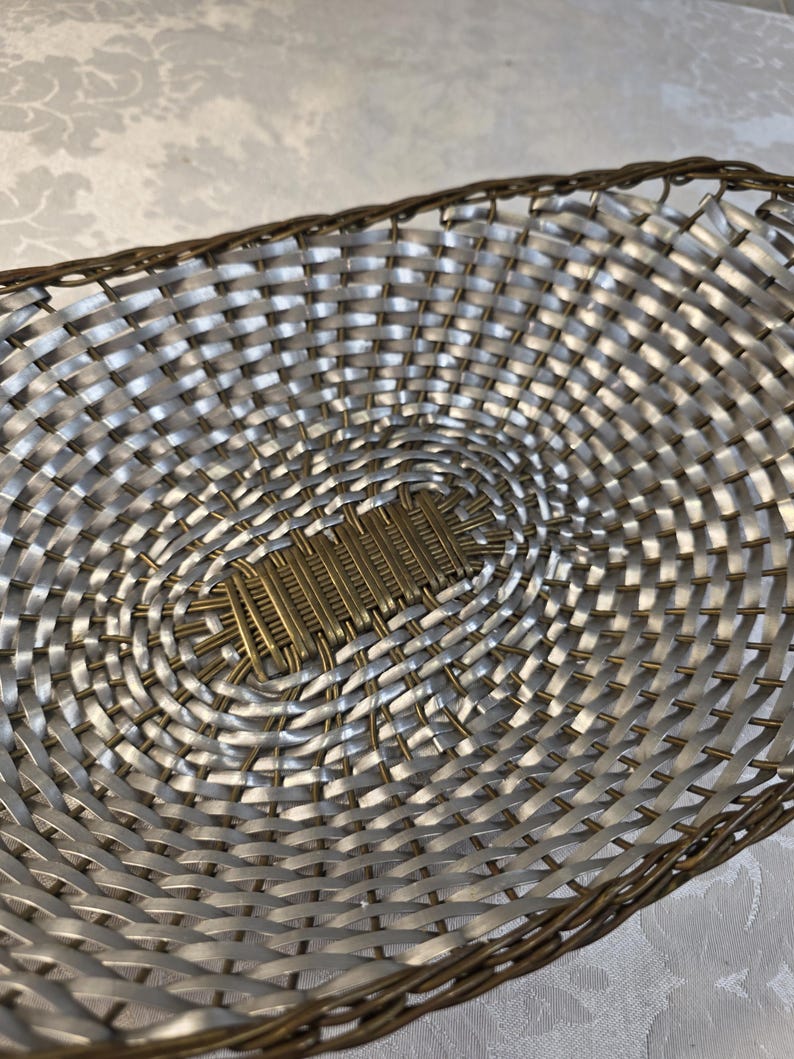 Puede incluir: Una cesta de forma ovalada con un patr&oacute;n trenzado de tiras plateadas met&aacute;licas y alambre fino de color bronce. La base central consiste en una secci&oacute;n rectangular de alambre de bronce tejido, mientras que el borde est&aacute; acabado en alambre.
