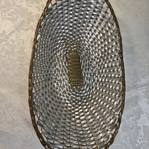 Puede incluir: Una cesta de forma ovalada con un dise&ntilde;o trenzado de tiras plateadas met&aacute;licas y un borde marr&oacute;n oscuro. La base de la cesta est&aacute; construida con una secci&oacute;n central rectangular de material marr&oacute;n oscuro, rodeada por el patr&oacute;n trenzado plateado.
