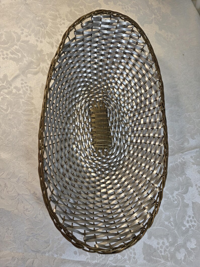 Puede incluir: Una cesta de forma ovalada con un dise&ntilde;o trenzado de tiras plateadas met&aacute;licas y un borde marr&oacute;n oscuro. La base de la cesta est&aacute; construida con una secci&oacute;n central rectangular de material marr&oacute;n oscuro, rodeada por el patr&oacute;n trenzado plateado.