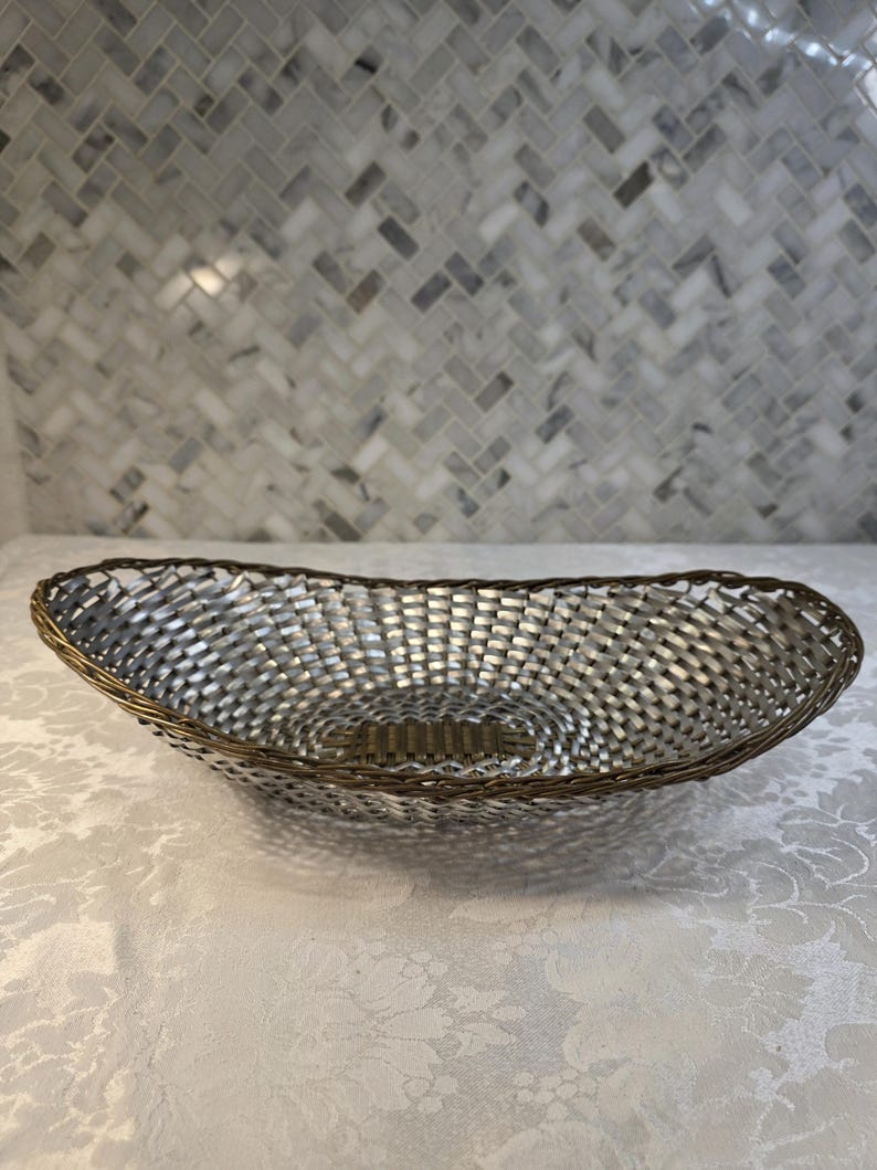 Puede incluir: Una cesta de forma ovalada con un cuerpo de metal plateado tejido, un borde de tono bronce oscuro y un detalle de base rectangular a juego. La estructura est&aacute; compuesta por finas tiras de metal entrelazadas que forman un recipiente abierto.