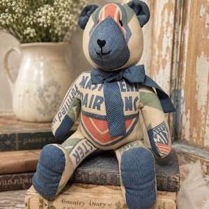 May include: A stuffed teddy bear constructed from repurposed fabric featuring blue corduroy accents on the ears, snout, and feet. The body displays printed text including MASTER MIX and AIR INC. A dark blue polka dot fabric bow is tied around the neck.