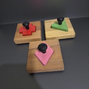 May include: Three wooden shape puzzles with black knobs. The shapes are a red cross, a green triangle, and a pink diamond, each set in a square wooden base. The background is a dark grey surface.