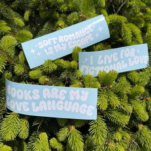 May include: Three light blue stickers with white text are displayed against a backdrop of green foliage. The stickers read: "SOFT ROMANCE IS MY ESCAPE", "I LIVE FOR FICTIONAL LOVE", and "BOOKS ARE MY LOVE LANGUAGE".