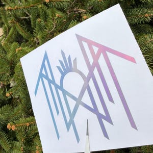 May include: A white paper with a geometric design in shades of blue, purple, and pink. The design features stylized arrows, a sun, and other geometric shapes. The paper is held against a backdrop of green pine needles.