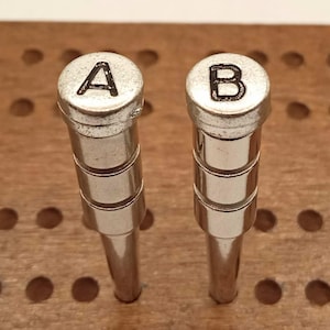 Puede incluir: Se muestran dos sellos de letras metálicas, uno con la letra "A" y otro con la letra "B". Los sellos tienen un acabado plateado y están colocados sobre un fondo de madera con agujeros espaciados uniformemente. Las letras son en negrita, en negro.