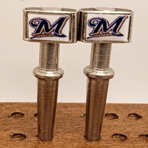 May include: Two metal cribbage pegs with a rectangular top featuring the letter "M" in blue and gold, set against a white background. The pegs are silver and are designed for use in a cribbage board.