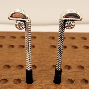 May include: Two silver-toned golf club-shaped objects with a golf ball detail, set against a wooden surface. The clubs have a textured, twisted design on the shaft and black bases. The golf balls are small and detailed.