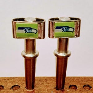 May include: Two metal bottle stoppers with a rectangular top featuring the Seattle Seahawks logo on a green background. The stoppers have a silver finish and a cylindrical stem. The stoppers are displayed on a wooden surface.