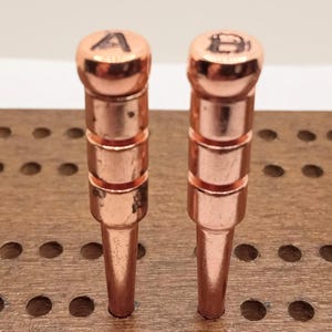 May include: Two copper-colored cribbage pegs with rounded tops, each with a letter, "A" and "B". The pegs are inserted into a wooden cribbage board. The pegs have a tiered design and a pointed base.