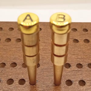 May include: Two gold-colored cribbage pegs with rounded tops, one marked with the letter "A" and the other with "B". The pegs are cylindrical with a tapered bottom, set in a wooden cribbage board.