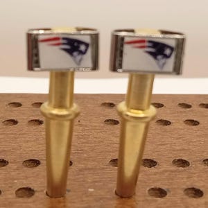May include: Two bottle stoppers with a rectangular top featuring the New England Patriots logo in red, white, and blue. The stoppers have a silver border and a gold-colored stem. They are displayed on a wooden surface.
